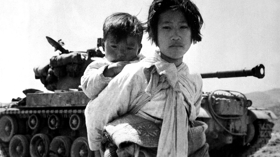 A photograph of a refugee carrying a baby in front of a tank